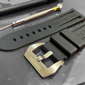 May include: Black rubber watch strap with a silver buckle, and the word "PANERAI" engraved on it. The strap has the word "OFFICINE" embossed on it. A black pen and a small screwdriver are also in the image.