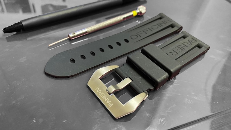 May include: Black rubber watch strap with a silver buckle, and the word "PANERAI" engraved on it. The strap has the word "OFFICINE" embossed on it. A black pen and a small screwdriver are also in the image.