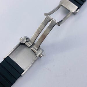 May include: A black rubber watch strap featuring a brushed stainless steel butterfly deployment clasp. The clasp is shown in an open position, revealing the metallic hinge mechanism and the textured black silicone band segments attached to it.