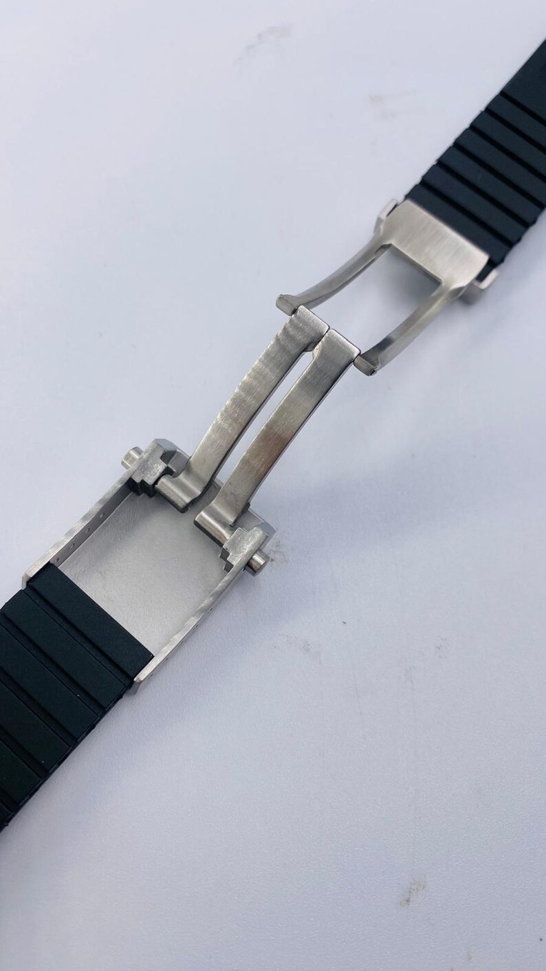May include: A black rubber watch strap featuring a brushed stainless steel butterfly deployment clasp. The clasp is shown in an open position, revealing the metallic hinge mechanism and the textured black silicone band segments attached to it.