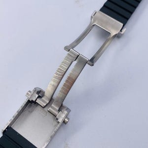 K&ouml;nnte beinhalten: Ein schwarzes Kautschuk-Uhrenarmband mit einer silberfarbenen Edelstahl-Faltschlie&szlig;e. Das Armband hat eine gerippte Textur und ist in einer offenen, ausgestreckten Position dargestellt, die den Metallscharniermechanismus zeigt.