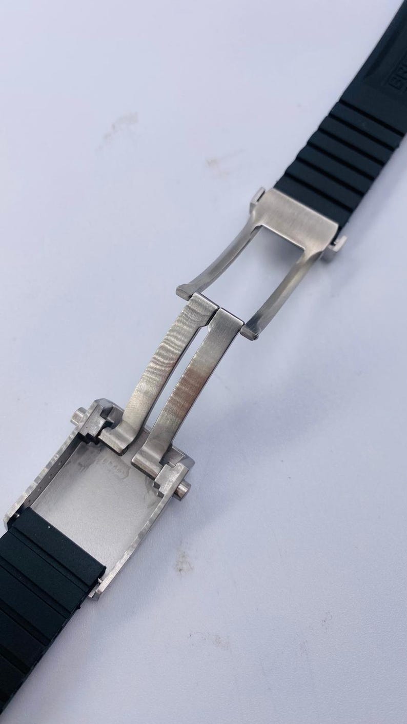 K&ouml;nnte beinhalten: Ein schwarzes Kautschuk-Uhrenarmband mit einer silberfarbenen Edelstahl-Faltschlie&szlig;e. Das Armband hat eine gerippte Textur und ist in einer offenen, ausgestreckten Position dargestellt, die den Metallscharniermechanismus zeigt.