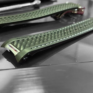 May include: Two olive green watch straps with a textured pattern and white stitching. The straps are lying on a reflective surface, with a small tool visible in the background. The watch straps are a replacement part.