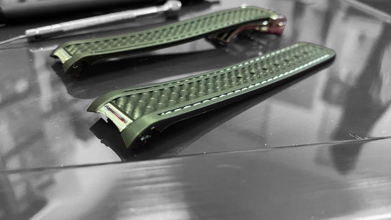 May include: Two olive green watch straps with a textured pattern and white stitching. The straps are lying on a reflective surface, with a small tool visible in the background. The watch straps are a replacement part.