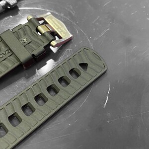 May include: Close-up of a watch strap and buckle. The strap is olive green with a textured pattern and rectangular cutouts. The buckle is gold-toned with a black strap holder. A metal tool is in the background.