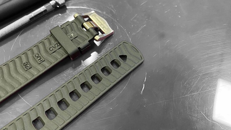 May include: Close-up of a watch strap and buckle. The strap is olive green with a textured pattern and rectangular cutouts. The buckle is gold-toned with a black strap holder. A metal tool is in the background.