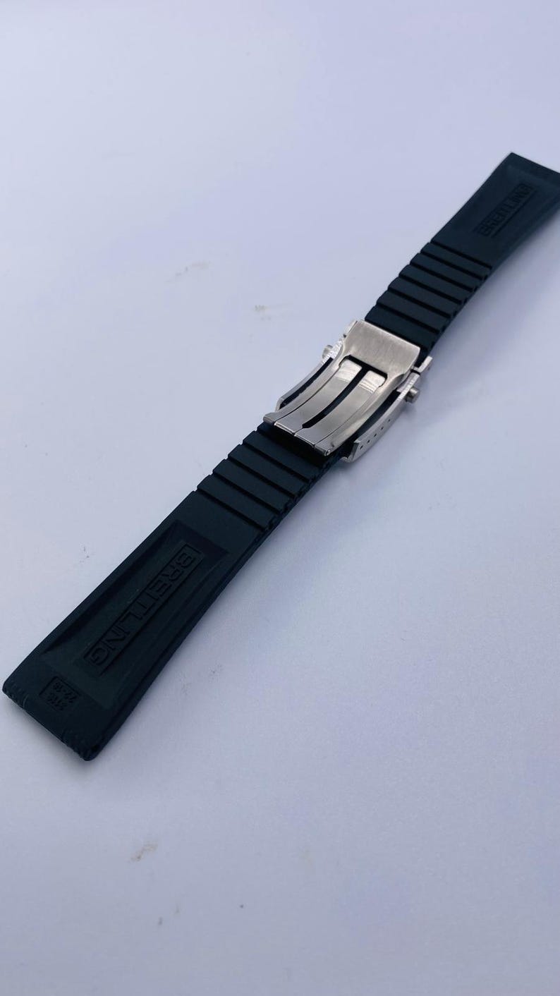 K&ouml;nnte beinhalten: Ein schwarzes Kautschuk-Uhrenarmband mit einer silberfarbenen Metall-Faltschlie&szlig;e. Das Armband hat eine gerippte Textur und das Wort BREITLING ist eingepr&auml;gt. Der Text 22-18 ist am Ende des Armbands deutlich zu erkennen.