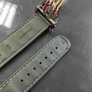 May include: Close-up of a watch strap in olive green leather with white stitching. The strap is marked with "CVZ007479" and "GJD 20". A portion of the watch clasp is visible, along with a small tool.