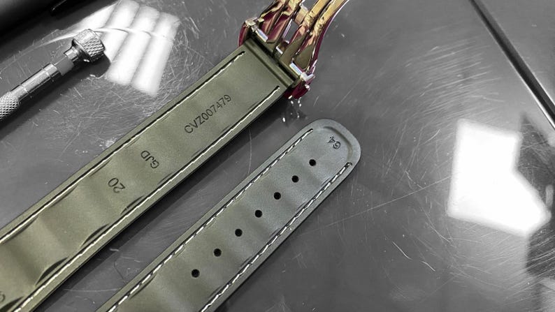 May include: Close-up of a watch strap in olive green leather with white stitching. The strap is marked with "CVZ007479" and "GJD 20". A portion of the watch clasp is visible, along with a small tool.
