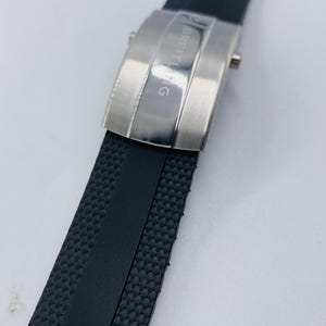 May include: A black rubber watch strap with a textured surface and a central smooth stripe. The strap features a brushed silver-tone metal deployment clasp with the word BREITLING engraved vertically on the surface of the buckle mechanism.