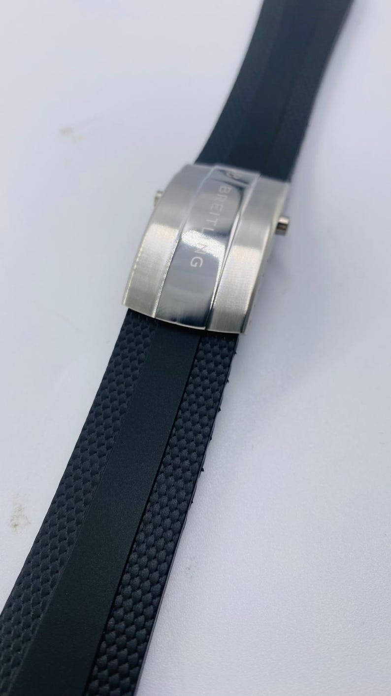 May include: A black rubber watch strap with a textured surface and a central smooth stripe. The strap features a brushed silver-tone metal deployment clasp with the word BREITLING engraved vertically on the surface of the buckle mechanism.