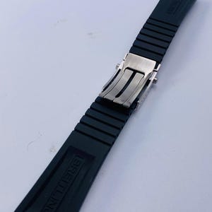 May include: A black rubber watch strap featuring a silver-toned metal deployment clasp. The strap has a ribbed texture and the word BREITLING is embossed on the surface of the rubber segments. The design includes a secure locking mechanism.