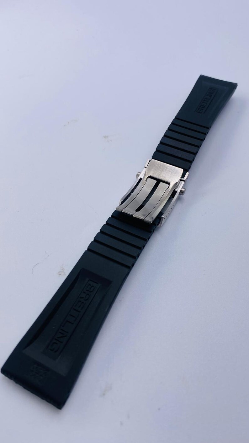 May include: A black rubber watch strap featuring a silver-toned metal deployment clasp. The strap has a ribbed texture and the word BREITLING is embossed on the surface of the rubber segments. The design includes a secure locking mechanism.