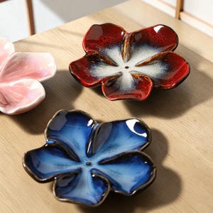 May include: Three ceramic flower-shaped dishes in shades of red, blue, and pink. The red dish has white and brown accents, the blue dish has a glossy finish, and the pink dish is partially visible. The dishes are on a light brown wooden surface.