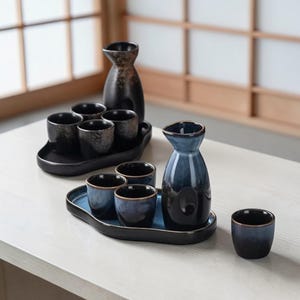 May include: Two sets of Japanese sake serving sets. Each set includes a ceramic carafe and four small cups on a tray. The carafes are dark, with one black and the other blue. The cups are dark blue and black.