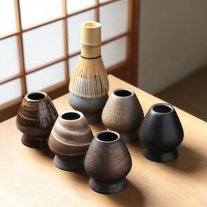 May include: A selection of ceramic matcha bowls in various earth-tone colours, arranged around a bamboo matcha whisk. The bowls have a unique, rounded shape and textured surfaces, ideal for a traditional tea ceremony.