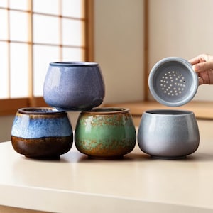 Może przedstawiać: Kolekcja ceramicznych filiżanek do herbaty w różnych kolorach, w tym niebieskim, zielonym i szarym. Jedna filiżanka ma pasującą pokrywkę z małymi otworami. Filiżanki mają zaokrąglony kształt i szkliwione wykończenie.