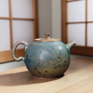 Puede incluir: Tetera redonda de cerámica con un esmalte azul-verdoso y un patrón moteado. La tetera tiene un asa curva, un pico y una tapa con un pequeño pomo. La tetera está sobre una superficie de madera.