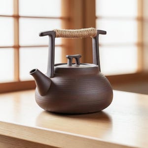 Könnte beinhalten: Eine braune Keramik-Teekanne mit einem Seilgriff. Die Teekanne hat einen Ausguss und einen Deckel mit einem kleinen Griff. Der Griff besteht aus einem dicken, naturfarbenen Seil. Die Teekanne steht auf einer Holzoberfläche.
