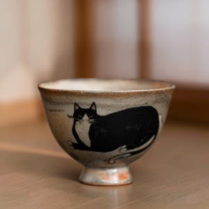 May include: A ceramic bowl with a black cat illustration. The bowl has a beige and brown color scheme with a pedestal base. The cat is lying down with its tail curled up. The bowl is likely used for serving food or as a decorative item.