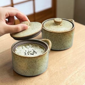 May include: Two speckled ceramic rice bowls with lids. One bowl is open, revealing white rice with black sesame seeds. The other bowl is closed. The bowls have small handles and a rustic, earthy aesthetic.