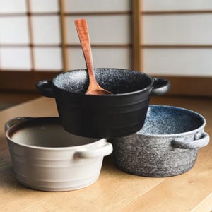 Puede incluir: Tres cuencos de cerámica en negro, gris y crema, cada uno con dos asas. Una cuchara de madera descansa en el cuenco negro. Los cuencos tienen una textura moteada y están dispuestos sobre una superficie de madera, lo que sugiere una cocina o un comedor.