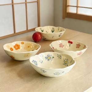 Può includere: Quattro ciotole in ceramica bianco sporco con bordi smerlati, ognuna decorata con un diverso motivo floreale in arancione, giallo, rosa e blu. Un frutto rosso è vicino alle ciotole. Le ciotole sono disposte su una superficie di legno chiaro.