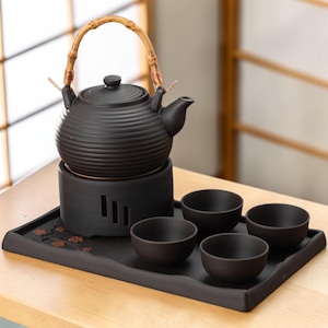May include: A black ceramic tea set with a bamboo handle on the teapot. The set includes a teapot, warmer, and four small teacups, all arranged on a rectangular tray. The teapot has a ribbed design.