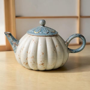 Puede incluir: Tetera de cerámica con cuerpo blanco y tapa azul. La tetera tiene forma redondeada con estrías verticales y un asa curva. La tapa está decorada con un motivo floral y un pequeño pomo. El pico y el asa tienen detalles azules y marrones.