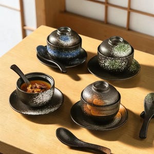 Może przedstawiać: Zestaw czterech ceramicznych misek do zupy w stylu japońskim z pokrywkami, spodkami i łyżkami. Miski są czarne z białymi plamkami i kolorowymi wzorami w kolorze niebieskim, zielonym i pomarańczowym. Łyżki i spodki są również czarne.