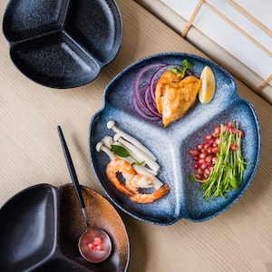 Könnte beinhalten: Dreigeteilte Keramikplatten in Dunkelblau und Schwarz. Eine Platte ist mit Essen gefüllt, darunter Garnelen, Pilze und Hähnchen. Ein schwarzer Löffel liegt auf einer ähnlichen Platte. Die Teller haben eine einzigartige, organische Form.