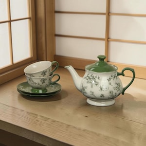 May include: A white ceramic tea set with a green floral pattern. The set includes a teapot, two teacups, and two saucers. The teapot has a green lid and handle. The teacups and saucers are stacked on a wooden surface.