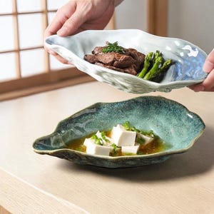 Può includere: Due piatti da portata in ceramica a forma di foglia. Un piatto contiene carne cotta e asparagi, l'altro cubetti di tofu in brodo, guarniti con cipollotti. I piatti hanno un design strutturato e organico con sfumature blu e verdi.