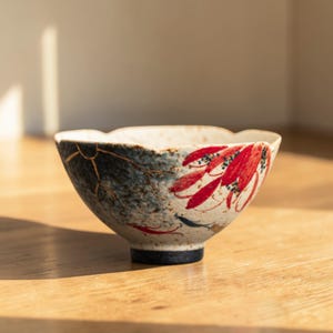 May include: A ceramic bowl with a unique design. The bowl features a blue and white exterior with red floral accents. The rim has a scalloped edge. The bowl has gold-colored kintsugi repair lines. The base is dark blue.