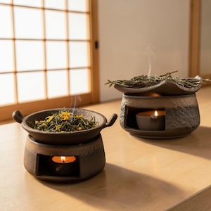 Op de afbeelding: Twee bruine keramische aromatherapie oliebranders met brandende kaarsen erin. Eén brander heeft een ronde schaal met handvatten, gevuld met groene kruiden en gele bloemknoppen. De andere brander heeft een blad-vormig deksel en is gevuld met kruiden.