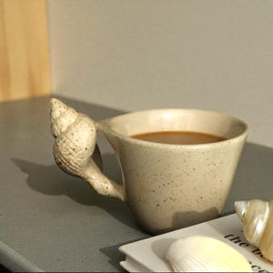Könnte beinhalten: Beige Keramiktasse mit einem muschelförmigen Griff. Die Tasse ist mit einer hellbraunen Flüssigkeit gefüllt, vermutlich Kaffee oder Tee. Die Tasse hat eine gesprenkelte Textur.