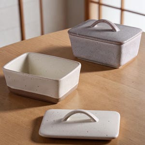 May include: Three ceramic butter dishes in a neutral color palette. One dish is open, revealing the interior, while another is closed with a lid and handle. The third lid is separate. The dishes have a speckled texture and a rectangular shape.