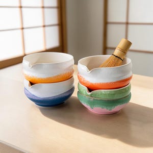 Puede incluir: Dos pilas de cuencos de matcha de cerámica colorida con un batidor de bambú. Cada cuenco es blanco en la parte superior, pasando a naranja, azul, verde y rosa. Los cuencos tienen un diseño de pico único para facilitar el vertido. El batidor es de bambú.