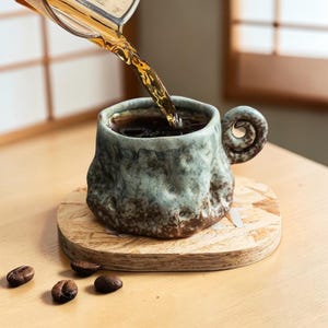 Puede incluir: Una taza de cerámica rústica, hecha a mano, con un exterior texturizado único y un asa en espiral. La taza se llena con líquido oscuro, posiblemente café, que se vierte de un recipiente de vidrio. Se apoya en un posavasos de madera, con granos de café.