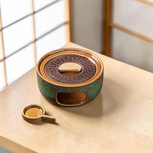 Può includere: Un scaldino per tè in ceramica con esterno verde e marrone e un top decorativo a motivi. Un piccolo piattino a forma di cucchiaio abbinato è posizionato accanto, su una superficie di legno chiaro. Lo scaldino ha un design circolare.