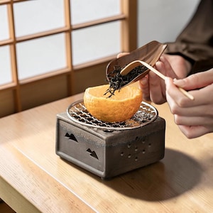 May include: A ceramic tea warmer with a metal grate holds a hollowed-out orange half. Tea leaves are being poured into the orange from a wooden scoop. The warmer has a dark, textured finish and mountain-shaped cutouts.