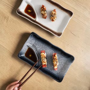 May include: Two rectangular sushi plates with a sauce compartment. Each plate holds two pieces of sushi topped with a creamy sauce and sesame seeds. One plate is white with a brown border, the other is gray. Chopsticks are near the gray plate.