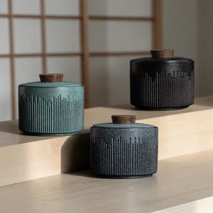 Puede incluir: Tres tarros de cerámica con tapas de madera. Los tarros son cilíndricos con líneas verticales texturizadas. Un tarro es verde azulado, otro negro y otro gris. Las tapas son de color madera oscura. Los tarros se exhiben sobre una superficie de madera clara.