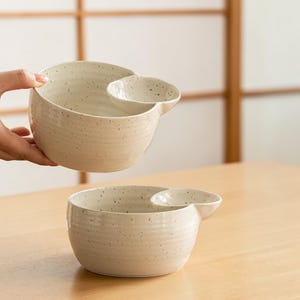 May include: Two off-white ceramic bowls with a speckled finish. Each bowl has a smaller, attached dish on the side. The bowls are on a light wooden surface. One bowl is being held by a hand.