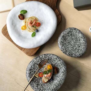 Könnte beinhalten: Ein weißer, unregelmäßig geformter Teller mit Spaghetti, Garnelen und Tomaten. Zwei gesprenkelte graue Teller und eine goldene Gabel sind ebenfalls zu sehen. Die Teller stehen auf einer Holzoberfläche.