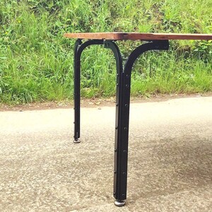 Könnte beinhalten: Holztisch mit dunkelbrauner Tischplatte und schwarzen Metallbeinen. Die Beine haben ein einzigartiges geschwungenes Design und sind mit sichtbaren Nieten an der Tischplatte befestigt. Der Tisch steht auf einer Betonoberfläche mit einem grasbewachsenen Hügel im Hintergrund.