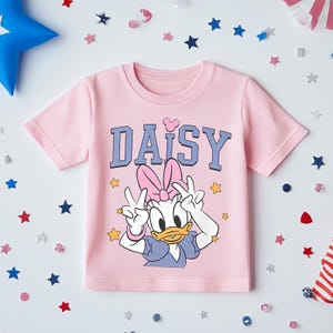 K&ouml;nnte beinhalten: Hellrosa T-Shirt mit dem Wort "DAISY" in blauen Buchstaben und einem Herz &uuml;ber dem "I". Das T-Shirt zeigt eine Grafik von Daisy Duck, die Friedenszeichen macht. Sternf&ouml;rmiges Konfetti umgibt das T-Shirt.