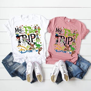 Puede incluir: Dos camisetas, una blanca y otra rosa, con el texto "My First Trip to Disney" y gr&aacute;ficos de personajes de dibujos animados. Las camisetas se combinan con vaqueros azules y zapatillas blancas, dispuestas sobre una superficie de madera blanca.