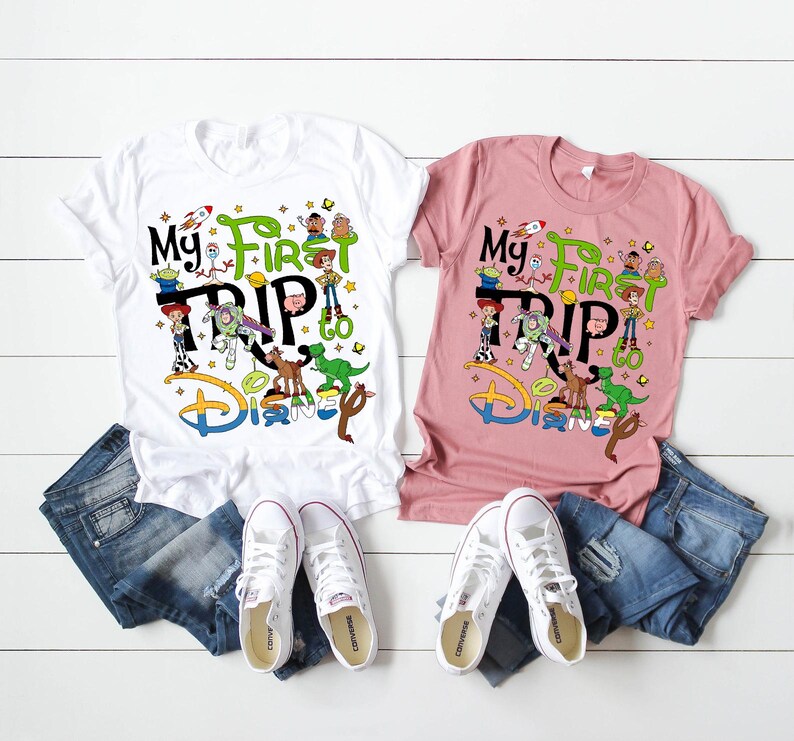 Puede incluir: Dos camisetas, una blanca y otra rosa, con el texto "My First Trip to Disney" y gr&aacute;ficos de personajes de dibujos animados. Las camisetas se combinan con vaqueros azules y zapatillas blancas, dispuestas sobre una superficie de madera blanca.