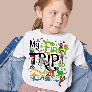 Puede incluir: Camiseta blanca con un gr&aacute;fico colorido con personajes de la pel&iacute;cula Toy Story y el texto "My First Trip to Disney". El dise&ntilde;o incluye un cohete, estrellas y el logotipo de Disney.